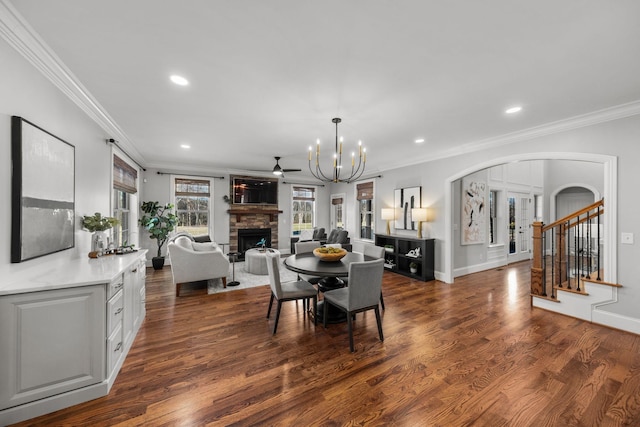 dining room featuring ceiling fan with notable chandelier, dark wood-type flooring, crown molding, and a stone fireplace