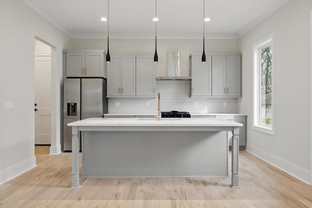 kitchen featuring a center island with sink, gray cabinetry, decorative backsplash, wall chimney range hood, and stainless steel fridge