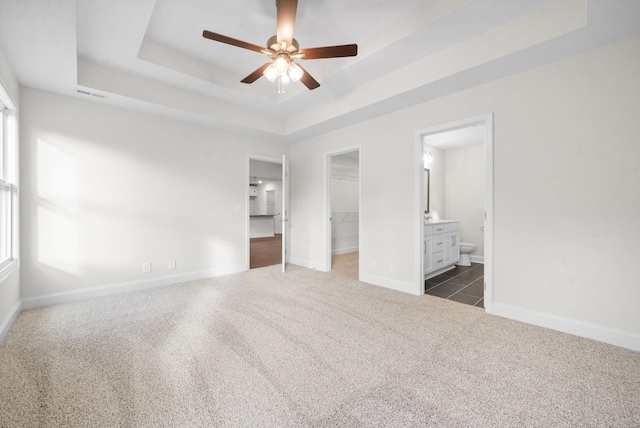 unfurnished bedroom featuring a spacious closet, dark colored carpet, a raised ceiling, ceiling fan, and ensuite bathroom