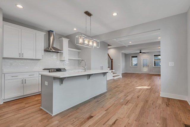 kitchen featuring ceiling fan, wall chimney range hood, white cabinets, and an island with sink