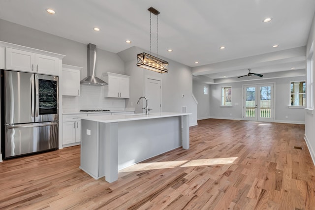 kitchen featuring an island with sink, stainless steel fridge, wall chimney range hood, and white cabinetry