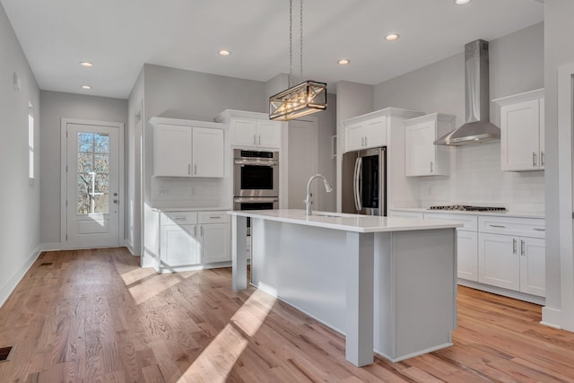 kitchen featuring white cabinetry, wall chimney range hood, stainless steel appliances, backsplash, and a kitchen island with sink