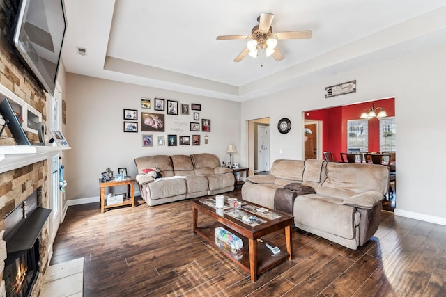 living room with a raised ceiling, dark hardwood / wood-style floors, ceiling fan with notable chandelier, and a stone fireplace