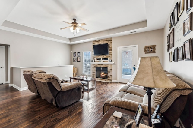 living room featuring dark wood-type flooring, a stone fireplace, a tray ceiling, and ceiling fan