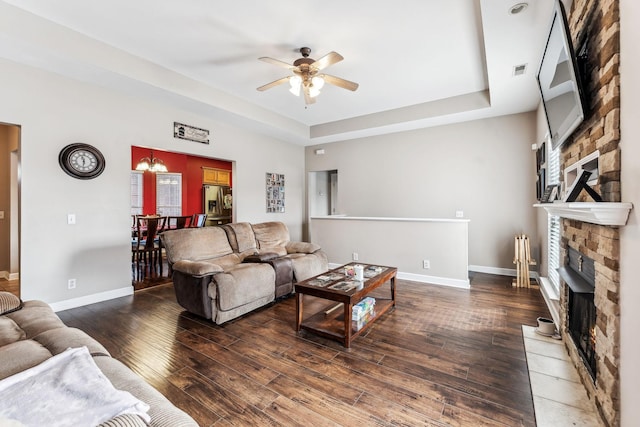 living room with ceiling fan with notable chandelier, dark wood-type flooring, a tray ceiling, and a stone fireplace