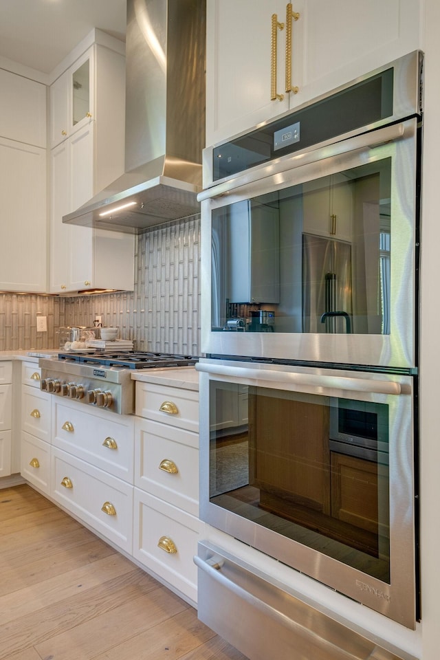 kitchen with white cabinetry, light hardwood / wood-style floors, stainless steel appliances, decorative backsplash, and wall chimney range hood