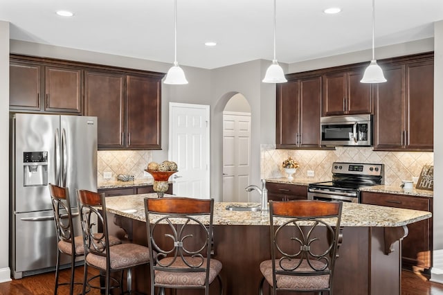 kitchen featuring a center island with sink, appliances with stainless steel finishes, dark brown cabinets, hanging light fixtures, and light stone countertops