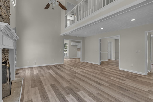 unfurnished living room with ceiling fan, a towering ceiling, light hardwood / wood-style flooring, a stone fireplace, and a textured ceiling