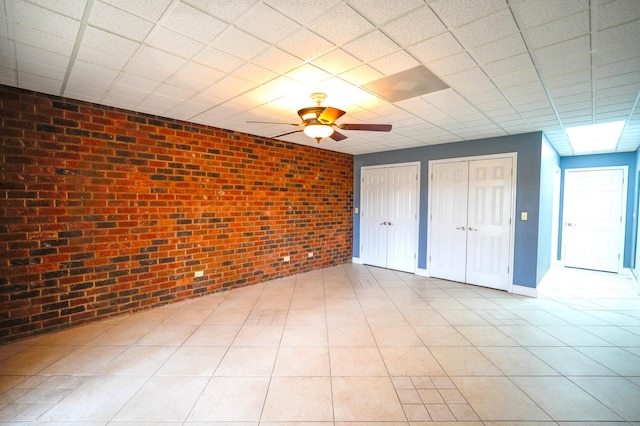 unfurnished bedroom featuring ceiling fan, two closets, brick wall, and a drop ceiling