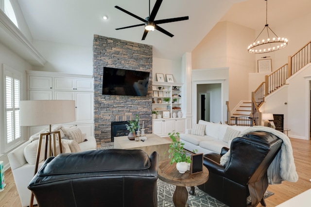 living room with a stone fireplace, high vaulted ceiling, ceiling fan with notable chandelier, light hardwood / wood-style flooring, and built in shelves