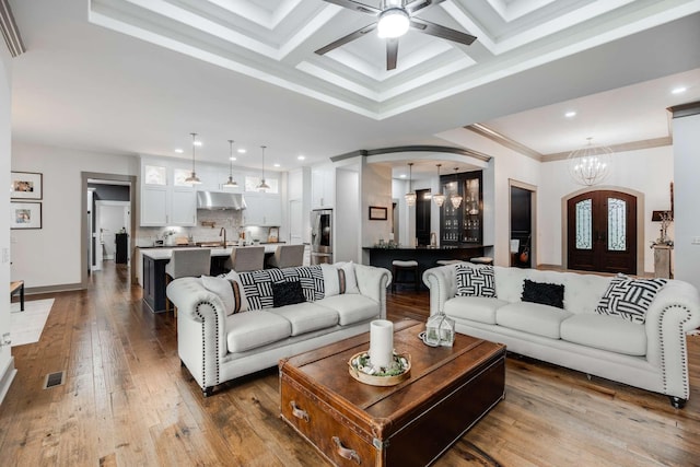 living room with beam ceiling, coffered ceiling, hardwood / wood-style flooring, ornamental molding, and ceiling fan with notable chandelier