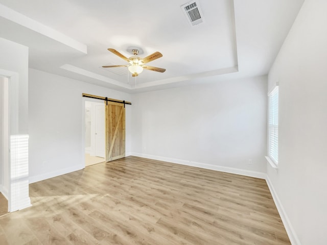 empty room with ceiling fan, a barn door, a tray ceiling, and light hardwood / wood-style flooring