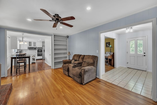 living room with ceiling fan with notable chandelier, built in shelves, ornamental molding, and light wood-type flooring