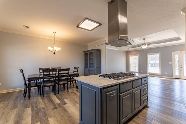 kitchen with ceiling fan with notable chandelier, a center island, island exhaust hood, stainless steel gas cooktop, and a tray ceiling