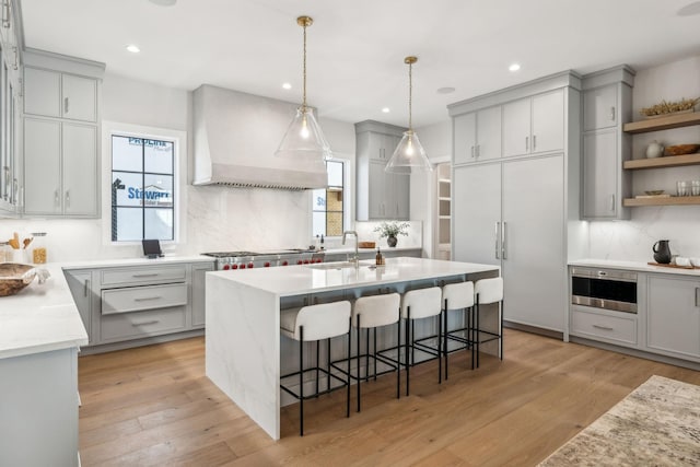 kitchen featuring a center island with sink, custom range hood, gray cabinetry, light wood-style floors, and a sink