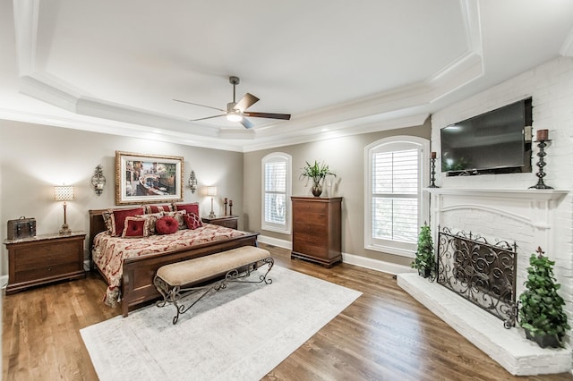 bedroom featuring a fireplace, hardwood / wood-style flooring, ornamental molding, ceiling fan, and a tray ceiling