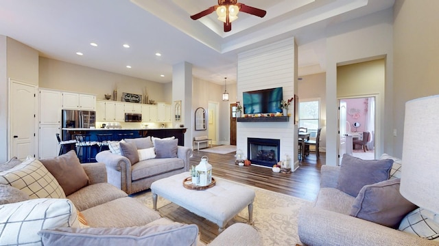 living room featuring ceiling fan, a raised ceiling, a towering ceiling, a large fireplace, and light wood-type flooring