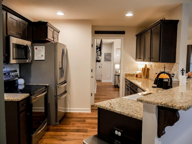 kitchen featuring appliances with stainless steel finishes, hardwood / wood-style flooring, kitchen peninsula, light stone counters, and dark brown cabinets