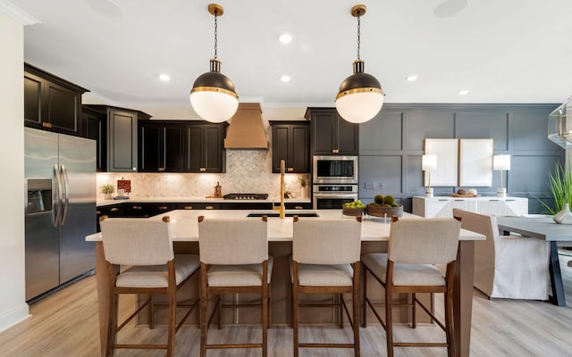 kitchen with premium range hood, stainless steel appliances, pendant lighting, and a center island with sink