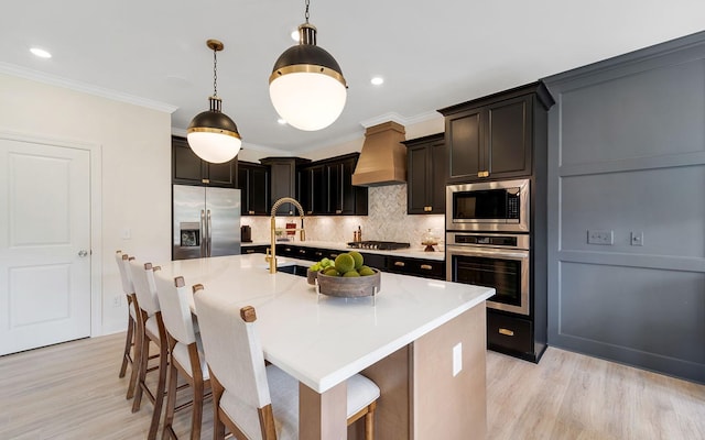 kitchen with a breakfast bar area, appliances with stainless steel finishes, a kitchen island with sink, custom range hood, and light hardwood / wood-style flooring