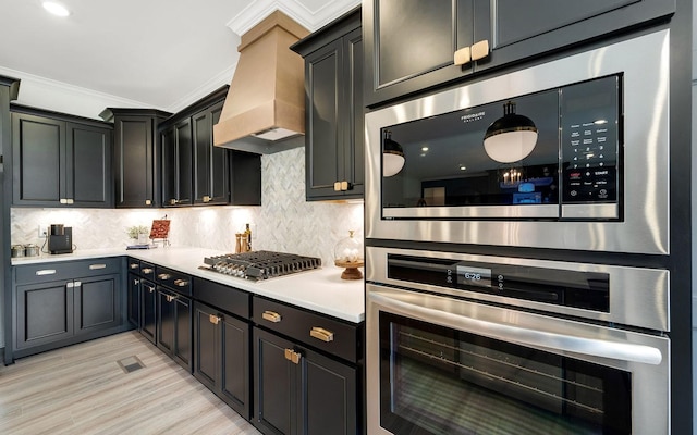 kitchen featuring custom range hood, stainless steel appliances, decorative backsplash, light hardwood / wood-style floors, and crown molding