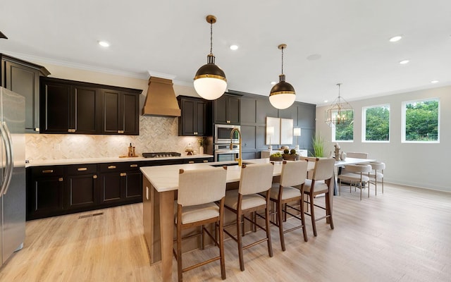 kitchen with custom exhaust hood, stainless steel appliances, hanging light fixtures, light wood-type flooring, and a center island with sink