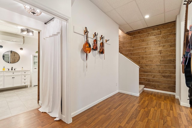 corridor with a wall mounted AC, sink, light wood-type flooring, wooden walls, and a drop ceiling