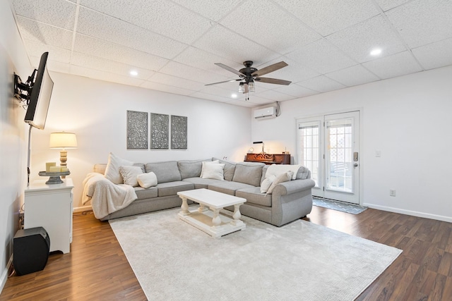 living room featuring an AC wall unit, dark wood-type flooring, a drop ceiling, and ceiling fan