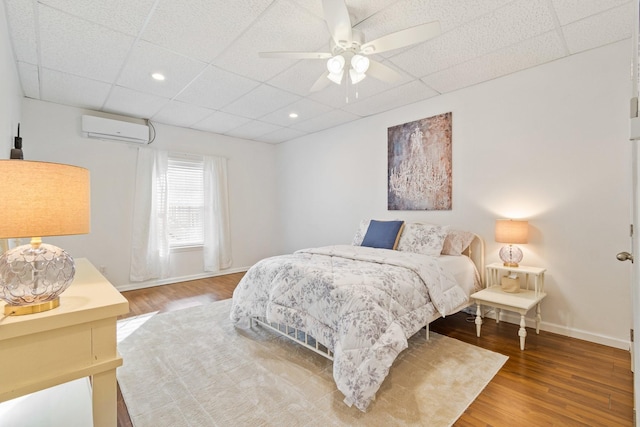 bedroom featuring ceiling fan, a paneled ceiling, hardwood / wood-style flooring, and a wall unit AC