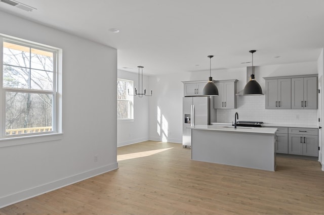 kitchen featuring tasteful backsplash, gray cabinets, stainless steel refrigerator with ice dispenser, hanging light fixtures, and a kitchen island with sink