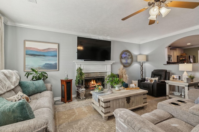 living room with crown molding, a textured ceiling, a stone fireplace, and ceiling fan