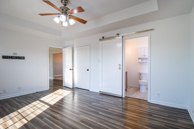 unfurnished bedroom featuring ensuite bath, a tray ceiling, a barn door, and dark hardwood / wood-style flooring