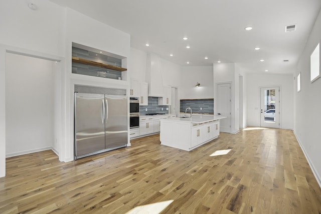 kitchen with appliances with stainless steel finishes, white cabinetry, decorative backsplash, light hardwood / wood-style floors, and a kitchen island with sink