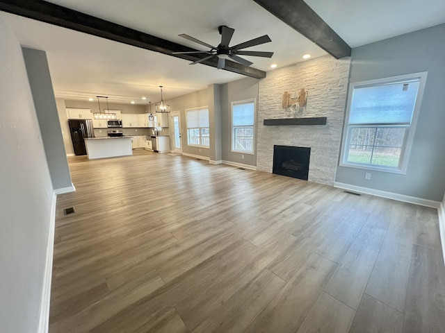 unfurnished living room with ceiling fan with notable chandelier, beamed ceiling, a stone fireplace, and light hardwood / wood-style floors