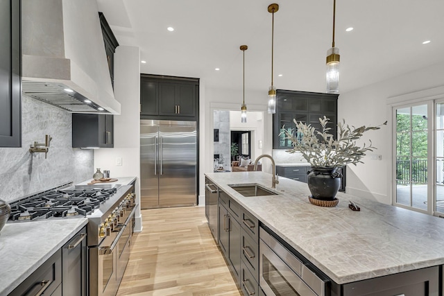 kitchen featuring an island with sink, hanging light fixtures, wall chimney range hood, and built in appliances