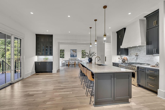 kitchen with backsplash, a large island with sink, light hardwood / wood-style flooring, high end stainless steel range oven, and wall chimney exhaust hood