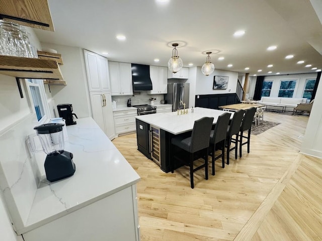 kitchen with pendant lighting, white cabinetry, wall chimney range hood, light stone counters, and a center island with sink