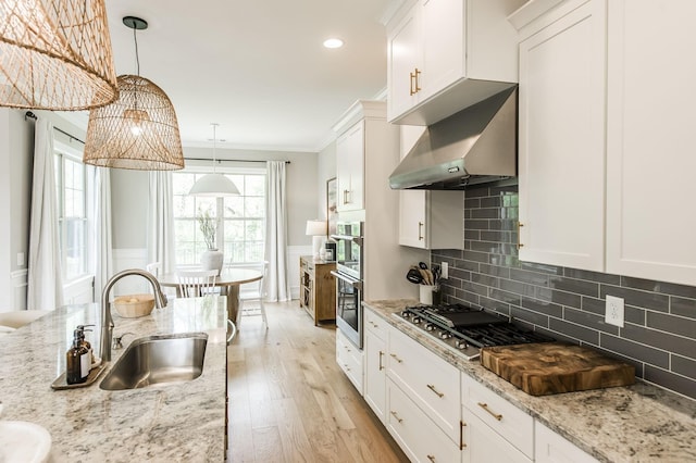 kitchen with stainless steel gas stovetop, wall chimney range hood, crown molding, white cabinetry, and hanging light fixtures