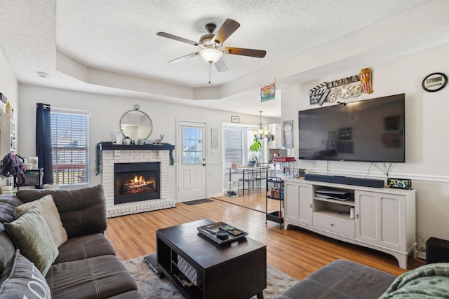 living room with ceiling fan with notable chandelier, a tray ceiling, light hardwood / wood-style flooring, and a brick fireplace