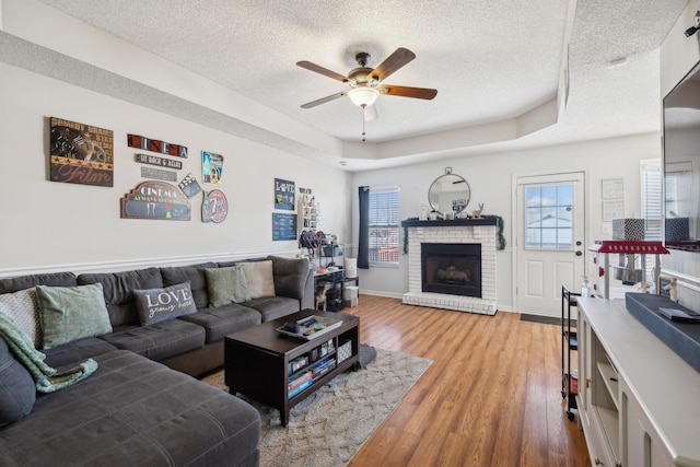 living room featuring hardwood / wood-style floors, a brick fireplace, ceiling fan, a raised ceiling, and a textured ceiling