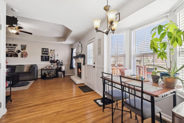 dining space featuring a raised ceiling, hardwood / wood-style flooring, a stone fireplace, a textured ceiling, and ceiling fan with notable chandelier