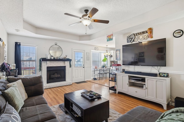 living room featuring a brick fireplace, light hardwood / wood-style floors, a tray ceiling, a textured ceiling, and ceiling fan with notable chandelier