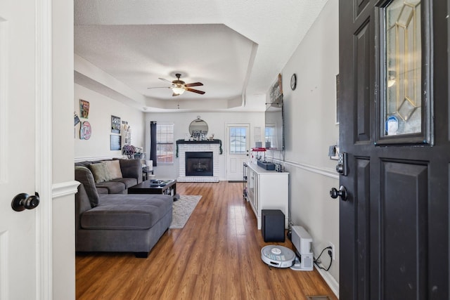 living room featuring ceiling fan, a raised ceiling, hardwood / wood-style flooring, a stone fireplace, and a textured ceiling