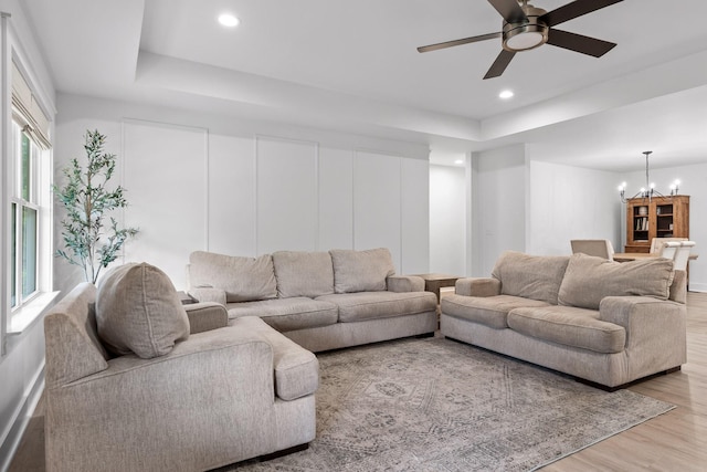 living area with a tray ceiling, light wood-type flooring, recessed lighting, and ceiling fan with notable chandelier