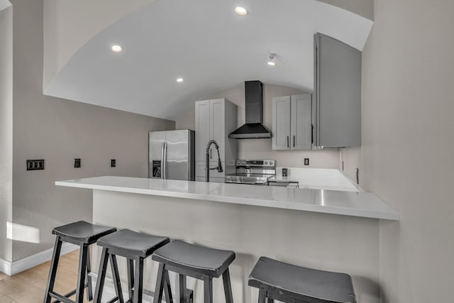 kitchen featuring kitchen peninsula, a breakfast bar area, appliances with stainless steel finishes, gray cabinetry, and wall chimney exhaust hood