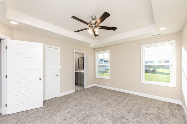 unfurnished bedroom featuring ceiling fan, connected bathroom, a tray ceiling, a closet, and light colored carpet