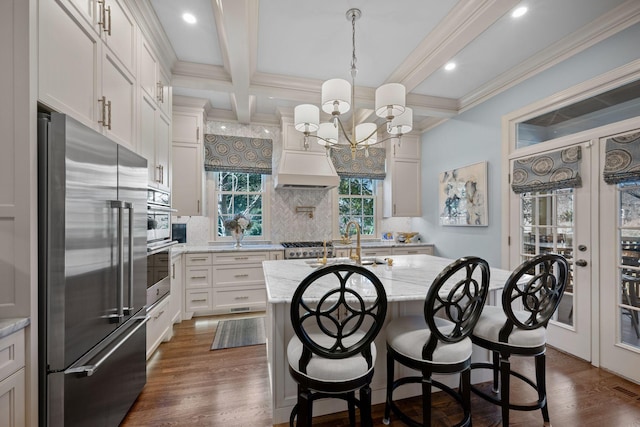 kitchen featuring light stone countertops, decorative light fixtures, stainless steel appliances, beamed ceiling, and a center island with sink