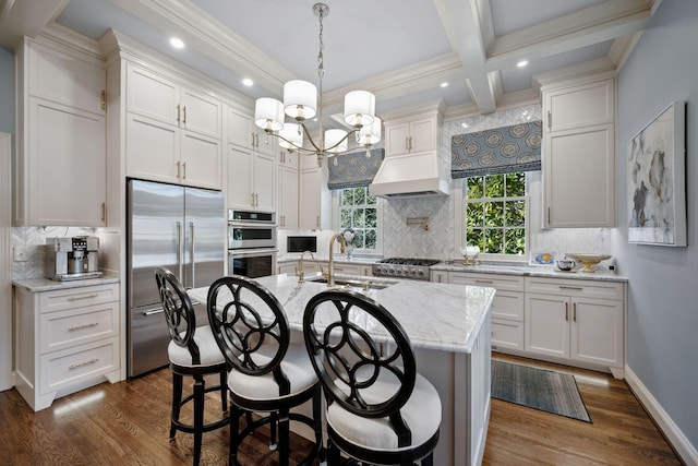 kitchen featuring an island with sink, stainless steel appliances, custom range hood, beam ceiling, and sink