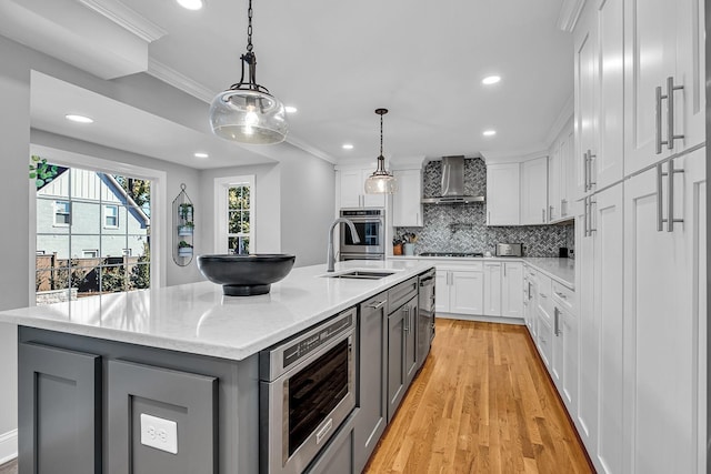 kitchen with wall chimney range hood, decorative backsplash, white cabinetry, appliances with stainless steel finishes, and an island with sink