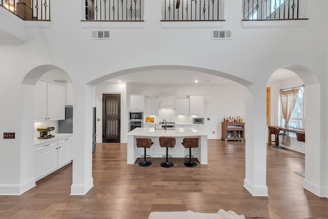 kitchen with a high ceiling, appliances with stainless steel finishes, white cabinetry, an island with sink, and dark hardwood / wood-style floors